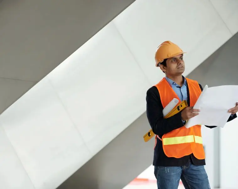 A man in an orange vest and hard hat holding papers.